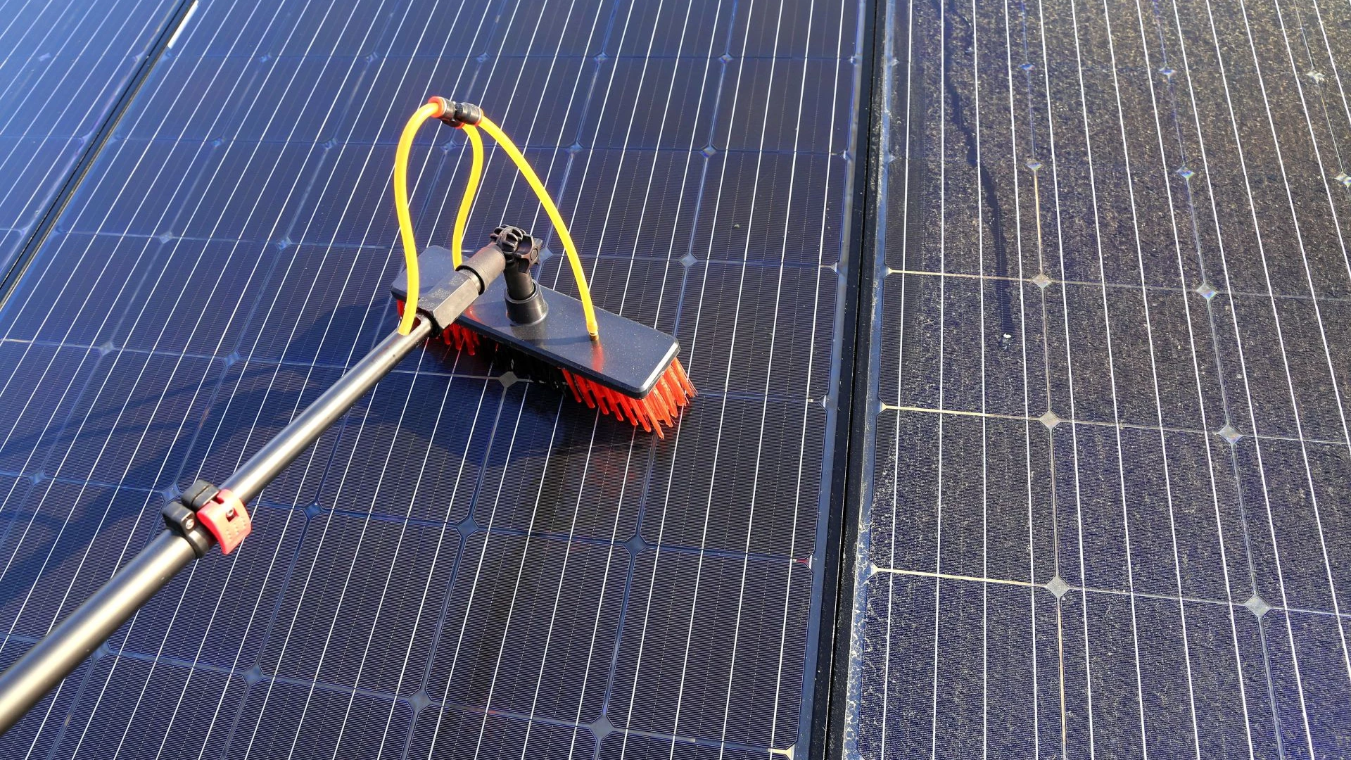Gros plan sur une brosse de nettoyage, fixée à une perche télescopique, lavant un panneau solaire bleu foncé avec de l'eau pure. La moitié gauche du panneau est propre et brillante, tandis que la moitié droite est encore couverte de poussière ou de saleté.